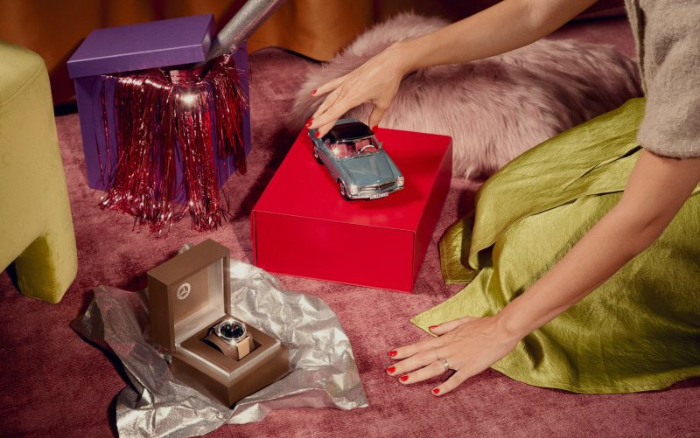 Mercedes-Benz Christmas Stars Zu sehen ist eine Geschenkeszene auf einem rosa Teppich.eine Person stellt ein Modellauto auf eine rote Geschenkbox. weitere Geschenkideen sind in Boxen zu sehen, darunter eine Armbanduhr.